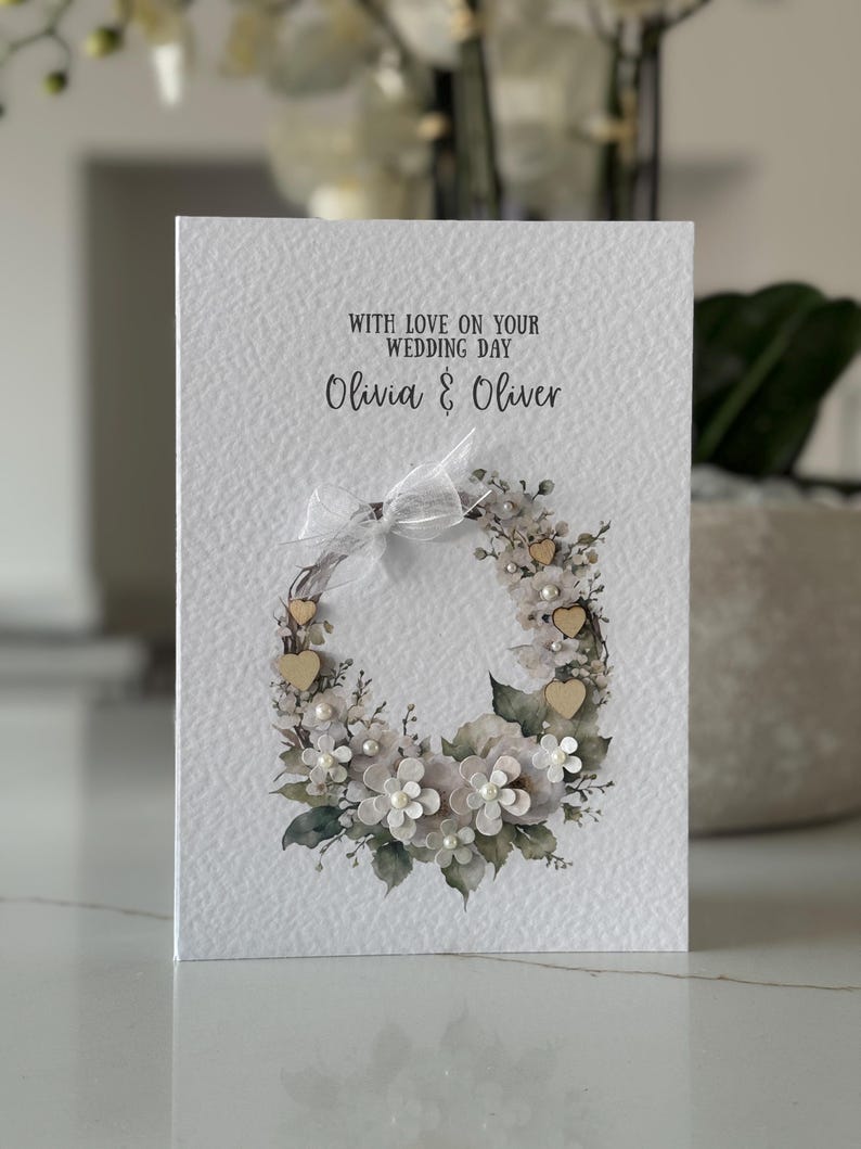 May include: A wedding card with a textured white background. The card features a floral wreath design with white flowers, pearls, and small wooden hearts. The text "WITH LOVE ON YOUR WEDDING DAY" and the names "Olivia & Oliver" are printed on the card.