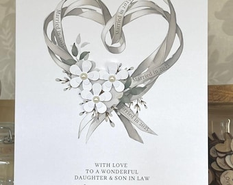 Könnte beinhalten: Hochzeitskarte mit einem herzförmigen Design aus grauen Bändern mit dem Text "Married in 2025". Weiße Blumen und Blätter befinden sich am unteren Rand. Die Karte enthält den Text "With love to a wonderful daughter & son in law, Olivia & Oscar, on your wedding day."