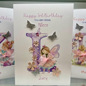 May include: A handmade birthday card with a purple letter "L" decorated with pink flowers and glitter. A fairy with pink wings and a pink dress is standing next to the letter. The card says "Happy 3rd Birthday to a very special Niece Lucy".