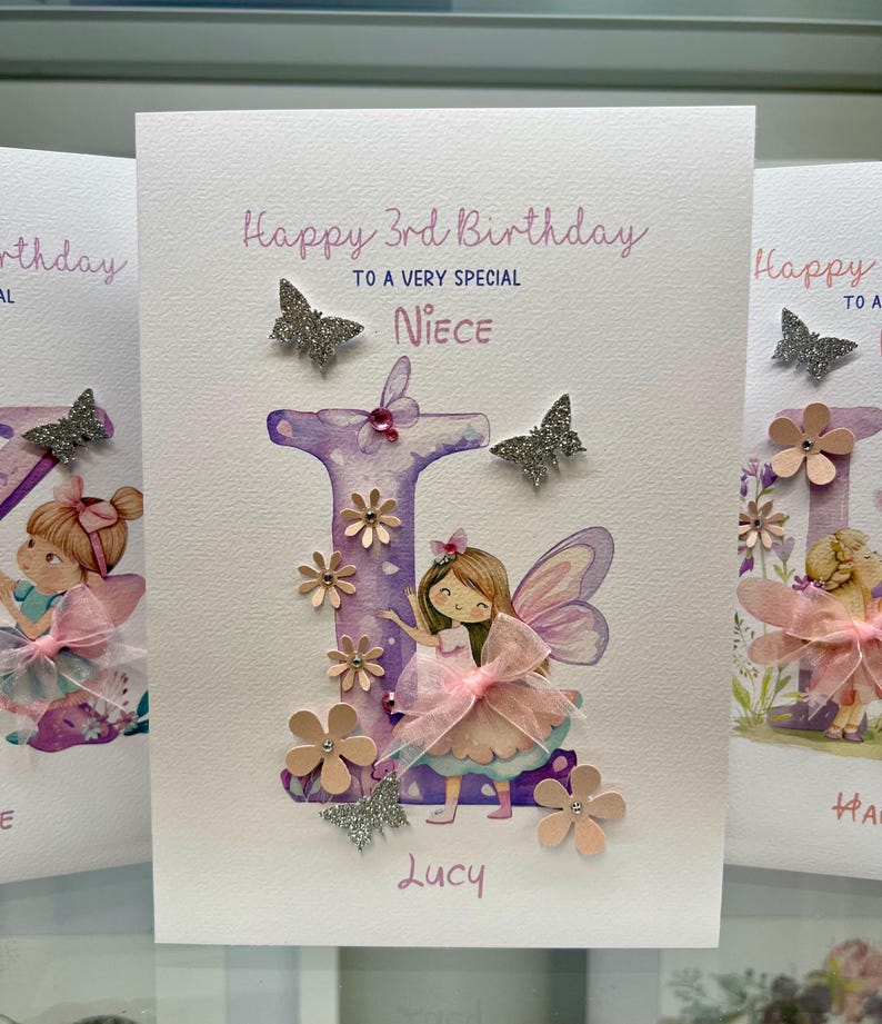 May include: A handmade birthday card with a purple letter "L" decorated with pink flowers and glitter. A fairy with pink wings and a pink dress is standing next to the letter. The card says "Happy 3rd Birthday to a very special Niece Lucy".
