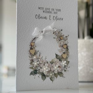 May include: A white wedding card with a floral wreath design. The card features the text "WITH LOVE ON YOUR WEDDING DAY" and the names "Olivia & Oliver". The wreath includes white flowers, pearls, and small wooden hearts.