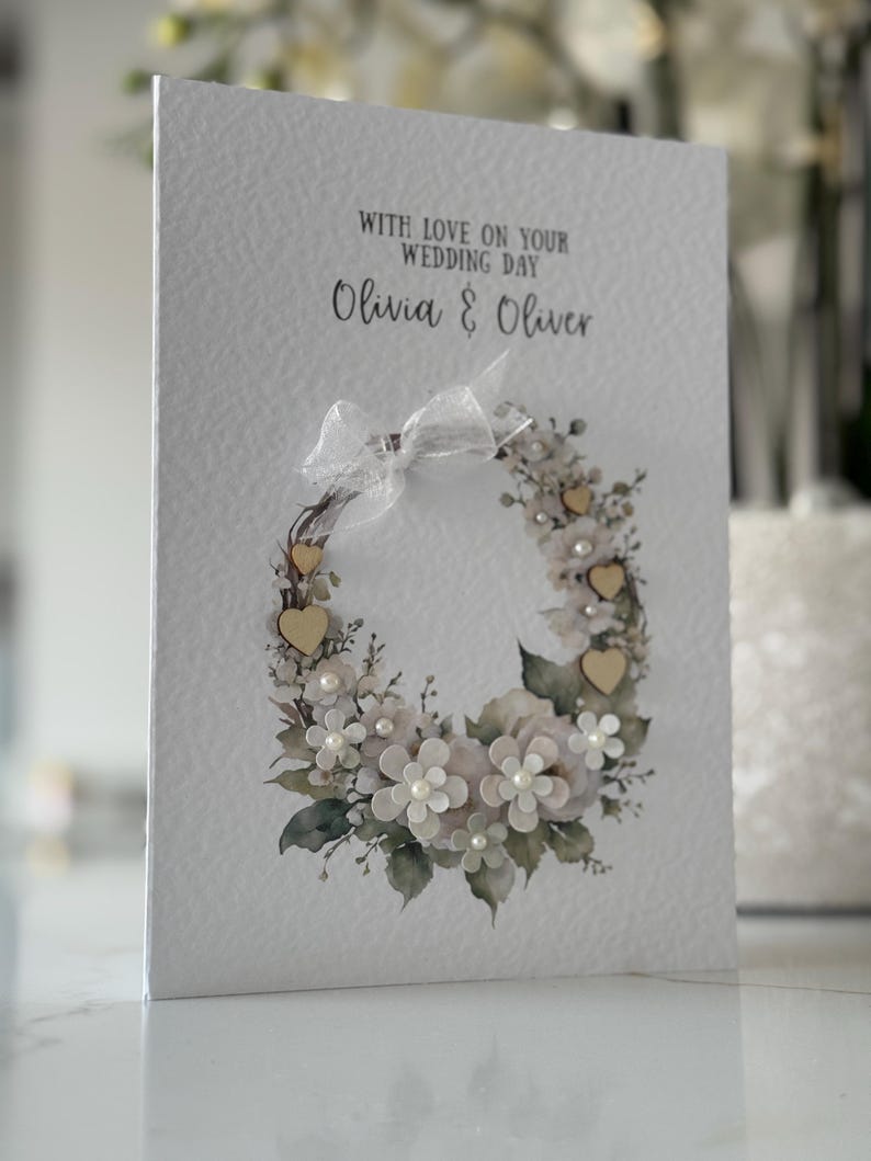 May include: A white wedding card with a floral wreath design. The card features the text "WITH LOVE ON YOUR WEDDING DAY" and the names "Olivia & Oliver". The wreath includes white flowers, pearls, and small wooden hearts.