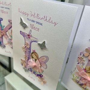 May include: A white card with the text "Happy 3rd Birthday to a very special Niece" and a pink and purple letter "L" with a fairy illustration. The letter is decorated with glitter and pink flowers.