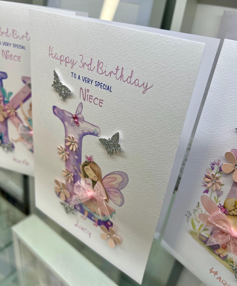 May include: A white card with the text "Happy 3rd Birthday to a very special Niece" and a pink and purple letter "L" with a fairy illustration. The letter is decorated with glitter and pink flowers.