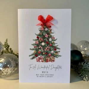 Puede incluir: Una tarjeta de Navidad con un diseño de árbol decorado. La tarjeta presenta un lazo rojo en la parte superior y un árbol de Navidad adornado con adornos y regalos. El texto dice: "To A Wonderful Daughter, Maya, May Your Christmas Be Merry & Bright."
