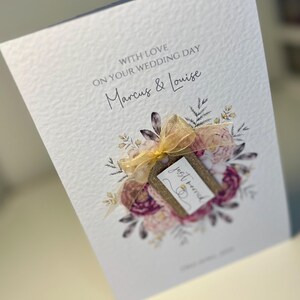 May include: A white card with a floral design and a gold ribbon. The card says "With love on your wedding day Marcus & Louise" and has a tag that says "Just married".