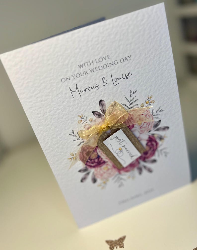 May include: A white card with a floral design and a gold ribbon. The card says "With love on your wedding day Marcus & Louise" and has a tag that says "Just married".