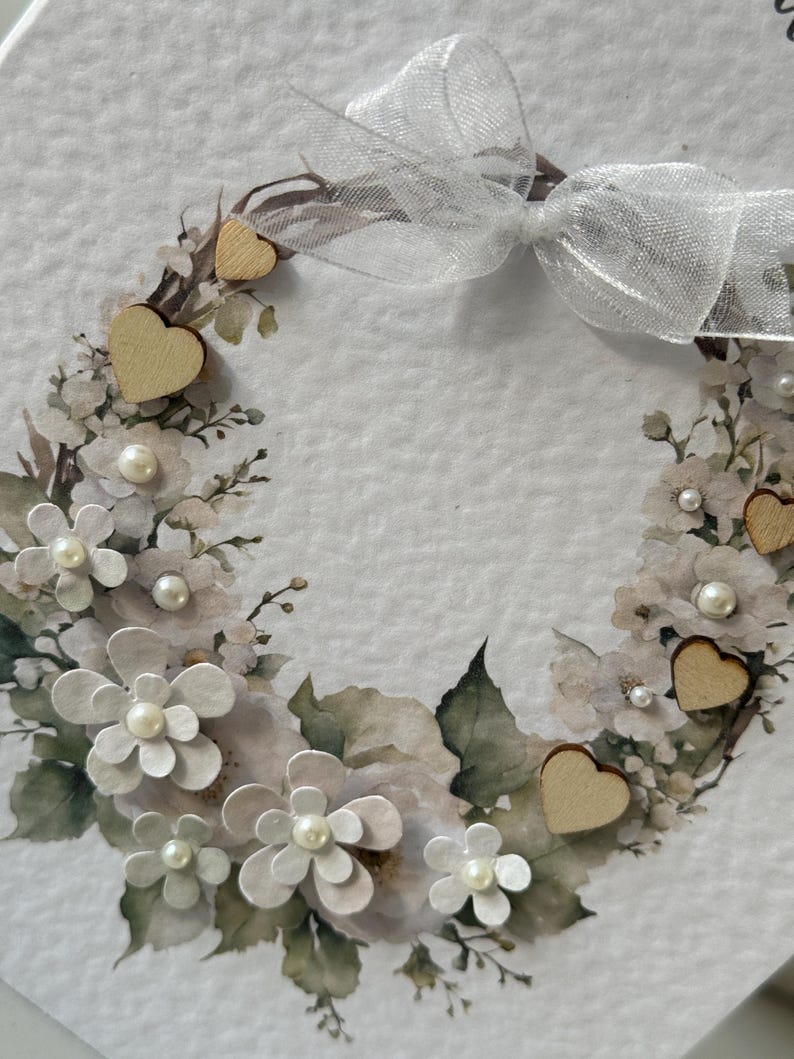 May include: A decorative wreath with white paper flowers, faux pearls, and small wooden hearts. A sheer white ribbon bow is at the top. The wreath is set against a white background.
