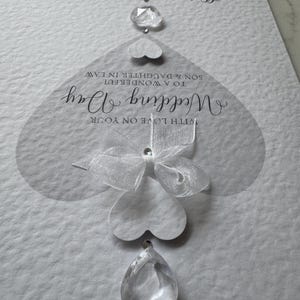 May include: A white wedding card with the words "Wedding Day" and "With Love on Your" in elegant script. The card features a series of heart-shaped embellishments and clear crystal accents, with the names "Oliver" and "Olivia" at the top.