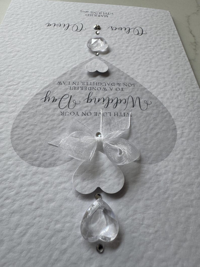 May include: A white wedding card with the words "Wedding Day" and "With Love on Your" in elegant script. The card features a series of heart-shaped embellishments and clear crystal accents, with the names "Oliver" and "Olivia" at the top.