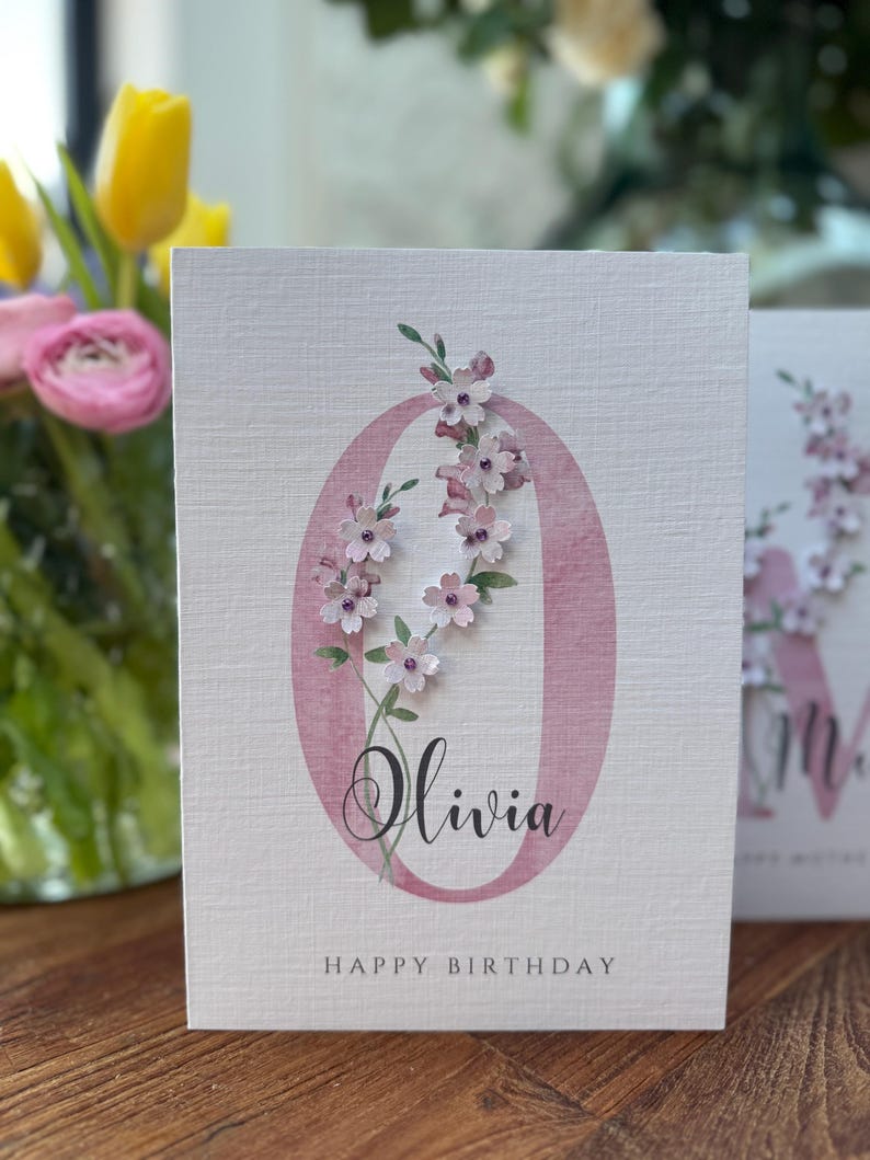 Peut inclure: Carte de v&oelig;ux blanche avec un "O" floral rose et le pr&eacute;nom "Olivia" en &eacute;criture cursive. La carte est orn&eacute;e de fleurs en papier et les mots "Happy Birthday" sont imprim&eacute;s en bas.