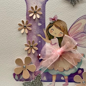 May include: A purple and pink fairy with brown hair and a pink bow is holding a pink ribbon. The fairy is standing in front of a purple letter I with pink flowers and glitter.