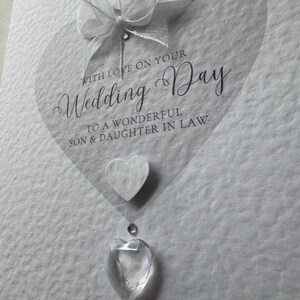 May include: A white wedding card with a transparent heart shape and the words "Wedding Day" in elegant script. Embellishments include a white bow, a felt heart, and clear crystal accents. The card reads "With Love on Your Wedding Day".