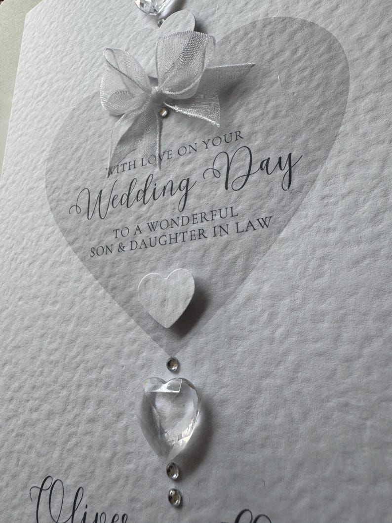May include: A white wedding card with a transparent heart shape and the words "Wedding Day" in elegant script. Embellishments include a white bow, a felt heart, and clear crystal accents. The card reads "With Love on Your Wedding Day".