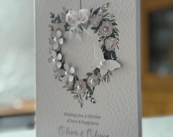 Könnte beinhalten: Eine Hochzeitskarte mit einem herzförmigen floralen Kranzdesign. Der Kranz zeigt weiße Blumen, Perlen und Grün. Auf der rechten Seite befindet sich ein weißer Schmetterling. Die Karte lautet "Wishing you a lifetime of love & happiness" und die Namen "Oliver & Olivia".