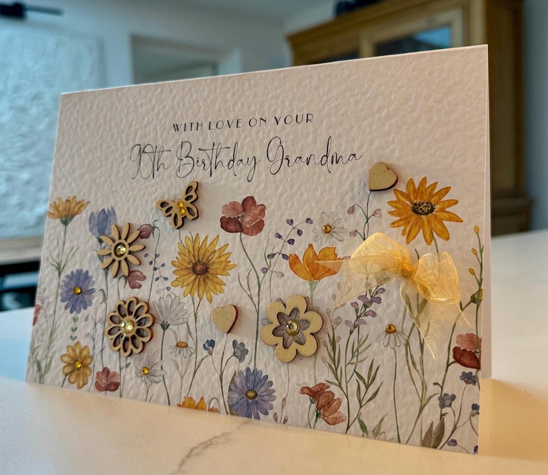 May include: A white card with a floral design and the text "With love on your 90th Birthday Grandma". The card has wooden embellishments in the shape of flowers, hearts, and a butterfly.