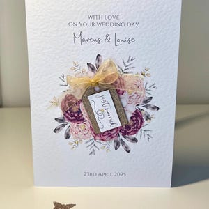 May include: A white wedding card with a floral design and a tag that says "just married". The card is decorated with gold glitter and a gold ribbon. The text on the card reads "With love on your wedding day Marcus & Louise 23rd April 2025".