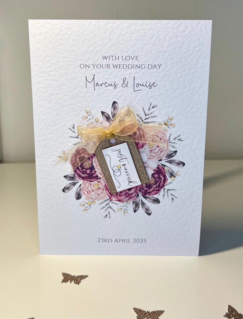 May include: A white wedding card with a floral design and a tag that says "just married". The card is decorated with gold glitter and a gold ribbon. The text on the card reads "With love on your wedding day Marcus & Louise 23rd April 2025".