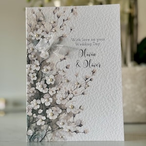 Może przedstawiać: Biała kartka ślubna z akwarelowym wzorem kwiatowym i perłowymi ozdobami. Na kartce widnieje napis "With love on your Wedding Day Olivia & Oliver".