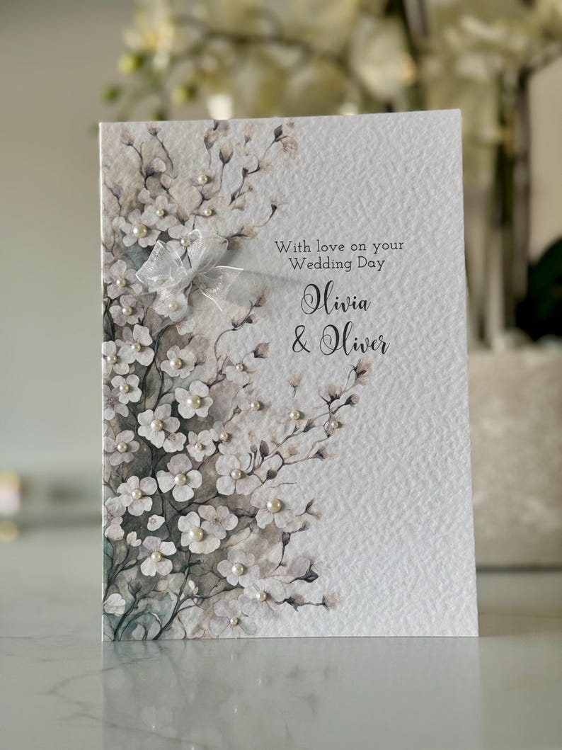 Może przedstawiać: Biała kartka ślubna z akwarelowym wzorem kwiatowym i perłowymi ozdobami. Na kartce widnieje napis "With love on your Wedding Day Olivia & Oliver".