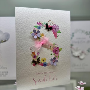May include: A white card with a pink and purple floral design featuring a large letter "S" with butterflies and the text "Happy Birthday Sarah Kate".