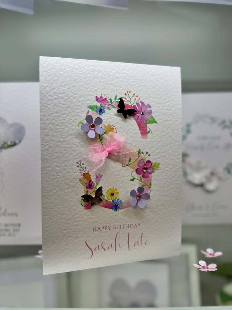 May include: A white card with a pink and purple floral design featuring a large letter "S" with butterflies and the text "Happy Birthday Sarah Kate".