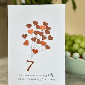 May include: White anniversary card featuring a heart-shaped bouquet in shades of brown and orange. The number 7 is displayed below the hearts, with the text "with love to my amazing Wife on our 7th Wedding Anniversary."