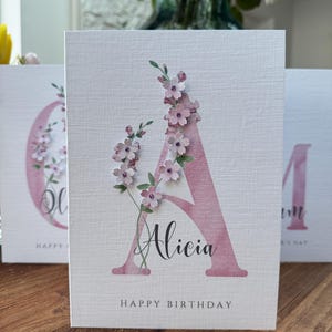 Peut inclure: Carte de v&oelig;ux avec un grand 'A' rose orn&eacute; de fleurs roses et de feuilles vertes. Le pr&eacute;nom 'Alicia' est &eacute;crit en script noir. Les mots 'Happy Birthday' sont imprim&eacute;s en bas.