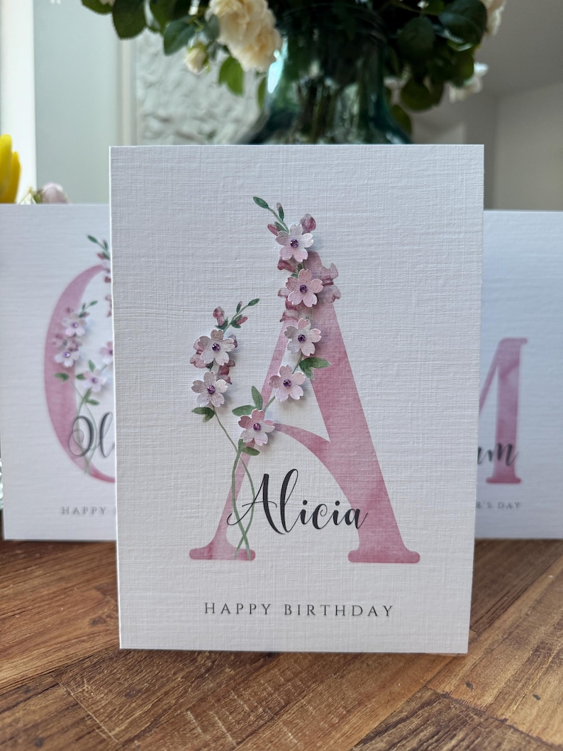 Peut inclure: Carte de v&oelig;ux avec un grand 'A' rose orn&eacute; de fleurs roses et de feuilles vertes. Le pr&eacute;nom 'Alicia' est &eacute;crit en script noir. Les mots 'Happy Birthday' sont imprim&eacute;s en bas.