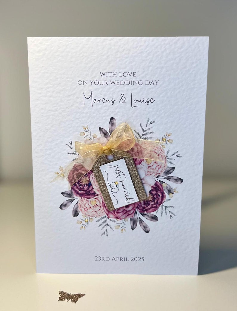 May include: A white wedding card with a floral wreath and a gold tag that says "just married". The card says "With love on your wedding day Marcus & Louise 23rd April 2025".