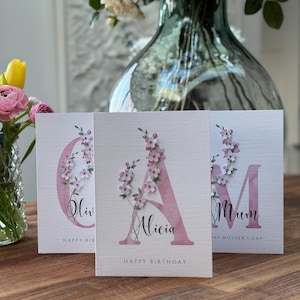 Peut inclure: Trois cartes de v&oelig;ux avec des motifs floraux. Les cartes pr&eacute;sentent les lettres "O", "A" et "M" en rose, avec des accents floraux. La carte du centre dit "Joyeux anniversaire" et le nom "Alicia". Les autres cartes disent "Bonne f&ecirc;te des m&egrave;res".