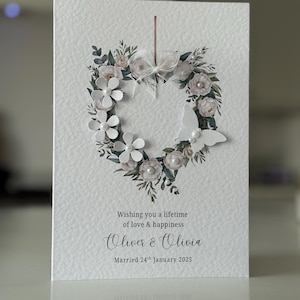May include: A white wedding card featuring a floral heart wreath. The design includes white paper flowers, pearls, and foliage. The card reads "Wishing you a lifetime of love & happiness", and the names "Oliver & Olivia", with a wedding date.