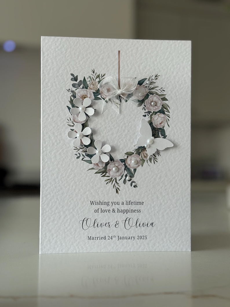 May include: A white wedding card featuring a floral heart wreath. The design includes white paper flowers, pearls, and foliage. The card reads "Wishing you a lifetime of love & happiness", and the names "Oliver & Olivia", with a wedding date.