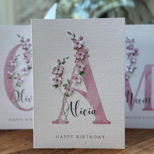 Peut inclure: Carte de v&oelig;ux textur&eacute;e avec un grand &laquo; A &raquo; rose orn&eacute; de d&eacute;licates fleurs roses. Le nom &laquo; Alicia &raquo; est &eacute;crit en dessous, avec &laquo; Happy Birthday &raquo; en bas. D'autres cartes sont visibles en arri&egrave;re-plan, avec le mot &laquo; Mum &raquo; visible.
