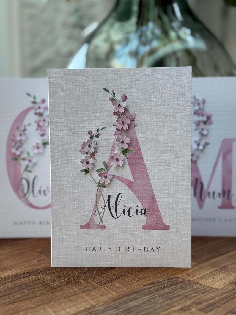 Peut inclure: Carte de v&oelig;ux textur&eacute;e avec un grand &laquo; A &raquo; rose orn&eacute; de d&eacute;licates fleurs roses. Le nom &laquo; Alicia &raquo; est &eacute;crit en dessous, avec &laquo; Happy Birthday &raquo; en bas. D'autres cartes sont visibles en arri&egrave;re-plan, avec le mot &laquo; Mum &raquo; visible.
