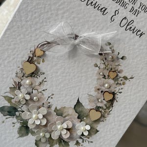 May include: A wedding card with a floral wreath design. The wreath features white paper flowers, faux pearls, and wooden heart accents. The card includes the text "With Love on Your Wedding Day" and the names "Olivia & Oliver". A sheer white ribbon bow is at the top.