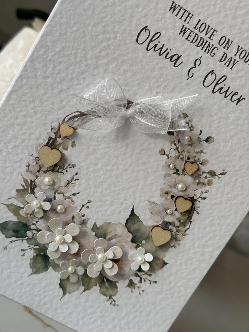 May include: A wedding card with a floral wreath design. The wreath features white paper flowers, faux pearls, and wooden heart accents. The card includes the text "With Love on Your Wedding Day" and the names "Olivia & Oliver". A sheer white ribbon bow is at the top.
