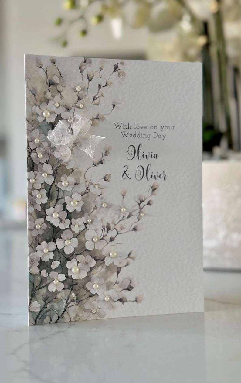 Może przedstawiać: Biała kartka ślubna z akwarelowym wzorem kwiatowym i perłowymi ozdobami. Na kartce widnieje napis "With love on your Wedding Day Olivia & Oliver".