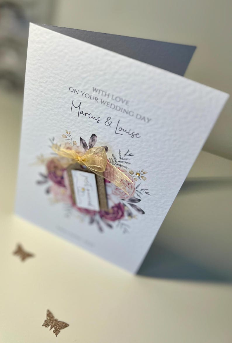 May include: A white wedding card with a floral design and a gold ribbon. The card says "With love on your wedding day Marcus & Louise".