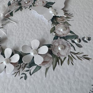 May include: A handmade greeting card with a floral wreath design. The wreath features white paper flowers, pearl accents, and a white butterfly. The card has a textured, off-white background.