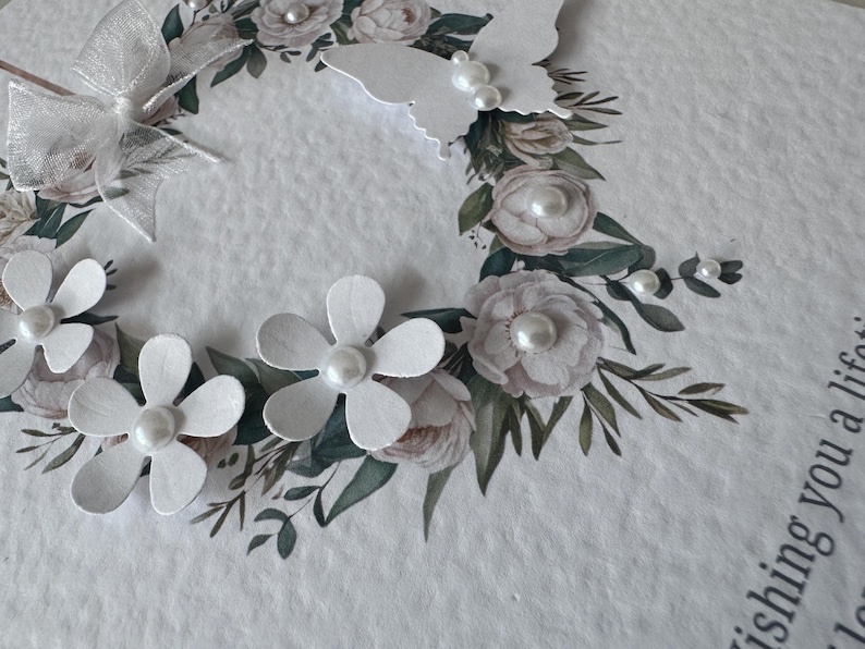 May include: A handmade greeting card with a floral wreath design. The wreath features white paper flowers, pearl accents, and a white butterfly. The card has a textured, off-white background.
