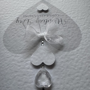 May include: A white wedding card with the words "Wedding Day" and "Congratulations" in elegant script. The card features a sheer heart-shaped overlay with a white bow and decorative crystal accents. A small heart and crystal pendant hang from the card.