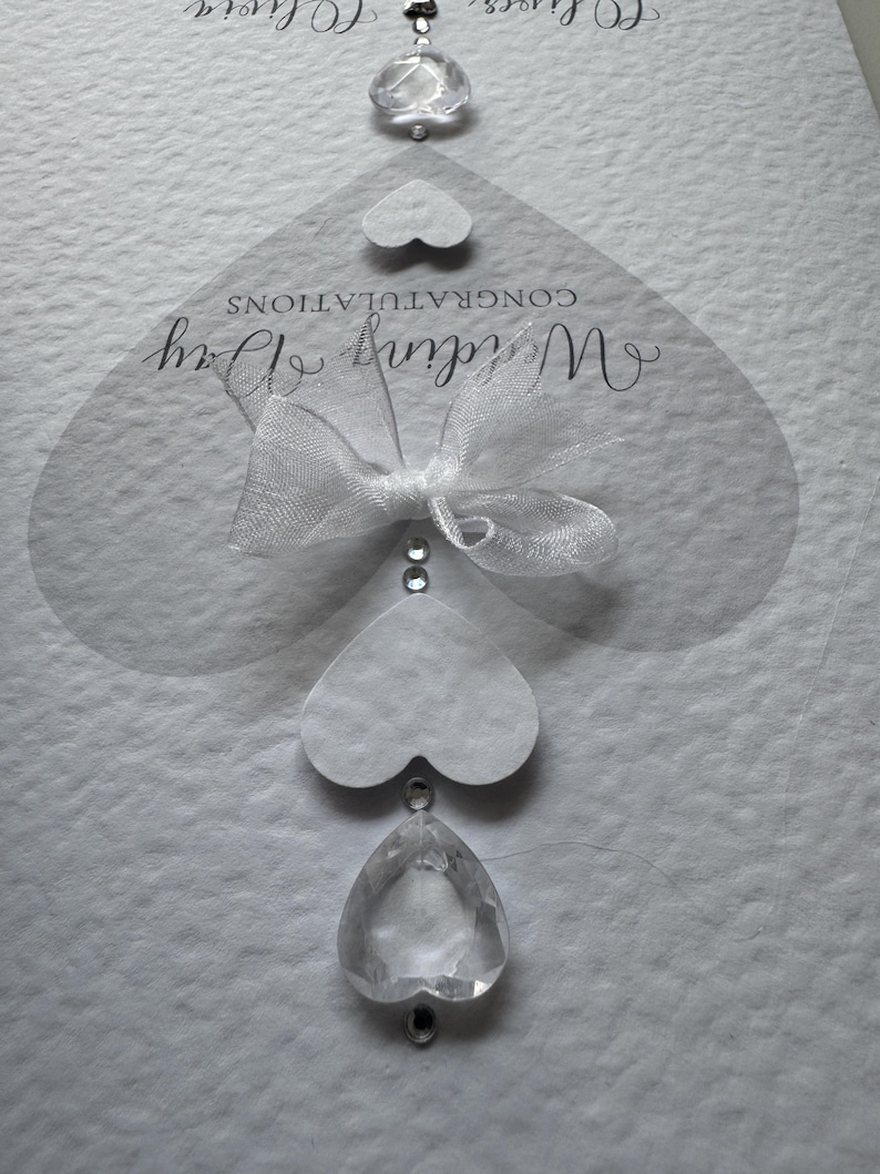 May include: A white wedding card with the words "Wedding Day" and "Congratulations" in elegant script. The card features a sheer heart-shaped overlay with a white bow and decorative crystal accents. A small heart and crystal pendant hang from the card.