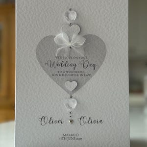 May include: A textured, light gray wedding card with a heart design. The card features the text "WITH LOVE ON YOUR Wedding Day" and the names "Oliver & Olivia" with the date "11th June 2025". Embellishments include clear heart-shaped gems and a white bow.