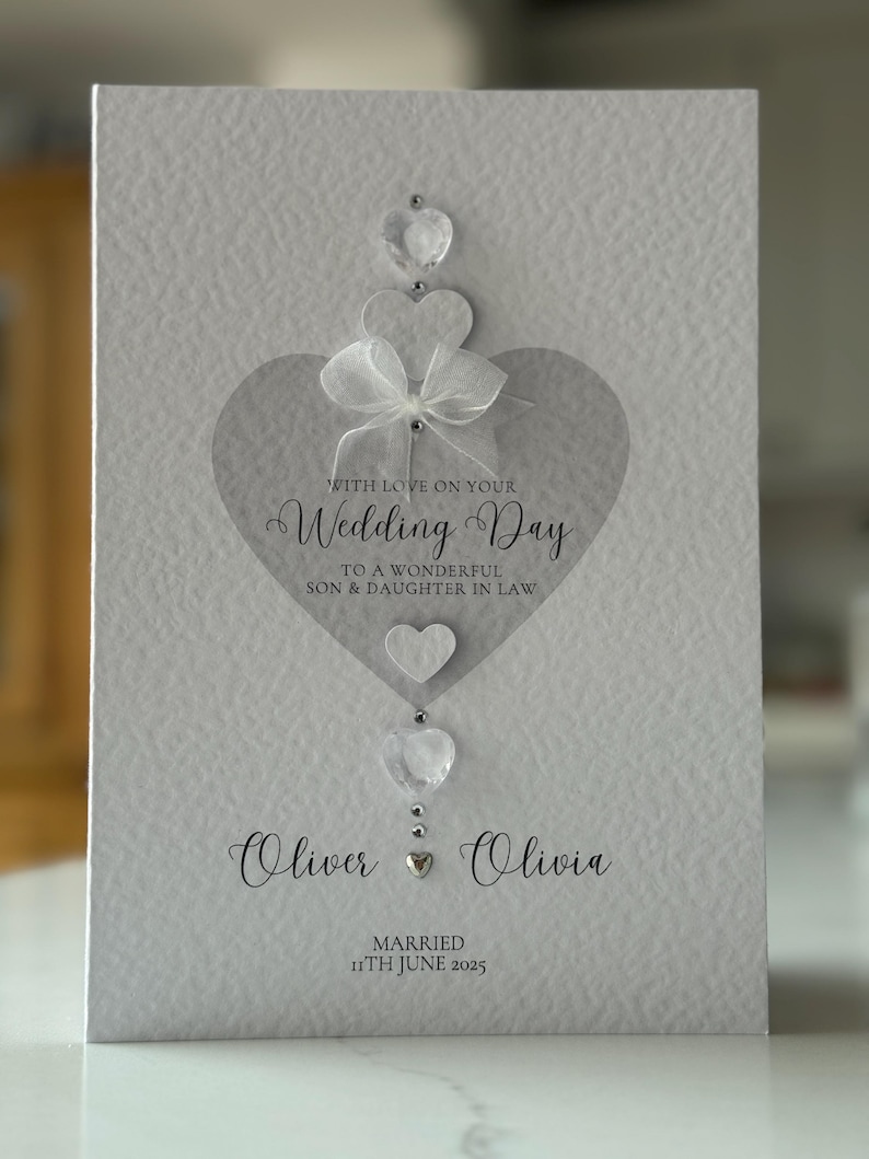 May include: A textured, light gray wedding card with a heart design. The card features the text "WITH LOVE ON YOUR Wedding Day" and the names "Oliver & Olivia" with the date "11th June 2025". Embellishments include clear heart-shaped gems and a white bow.