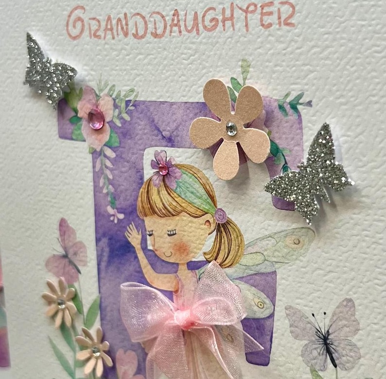 May include: A handmade greeting card with a purple letter "F" and a fairy illustration. The card is decorated with glitter butterflies, flowers, and rhinestones. The text "Granddaughter" is written at the top of the card.