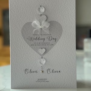 May include: A white wedding card with a heart design and the words "Wedding Day" in elegant script. The card includes the names "Oliver & Olivia" and the date "11th June 2025". Embellished with clear gems and a white bow.