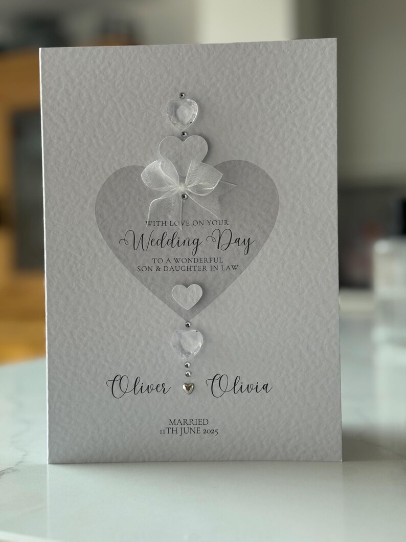 May include: A white wedding card with a heart design and the words "Wedding Day" in elegant script. The card includes the names "Oliver & Olivia" and the date "11th June 2025". Embellished with clear gems and a white bow.