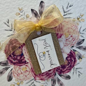 May include: A gold glitter tag with a white card that says "just married" in black calligraphy. The tag is tied with a gold ribbon and is surrounded by a floral arrangement of pink and purple flowers.