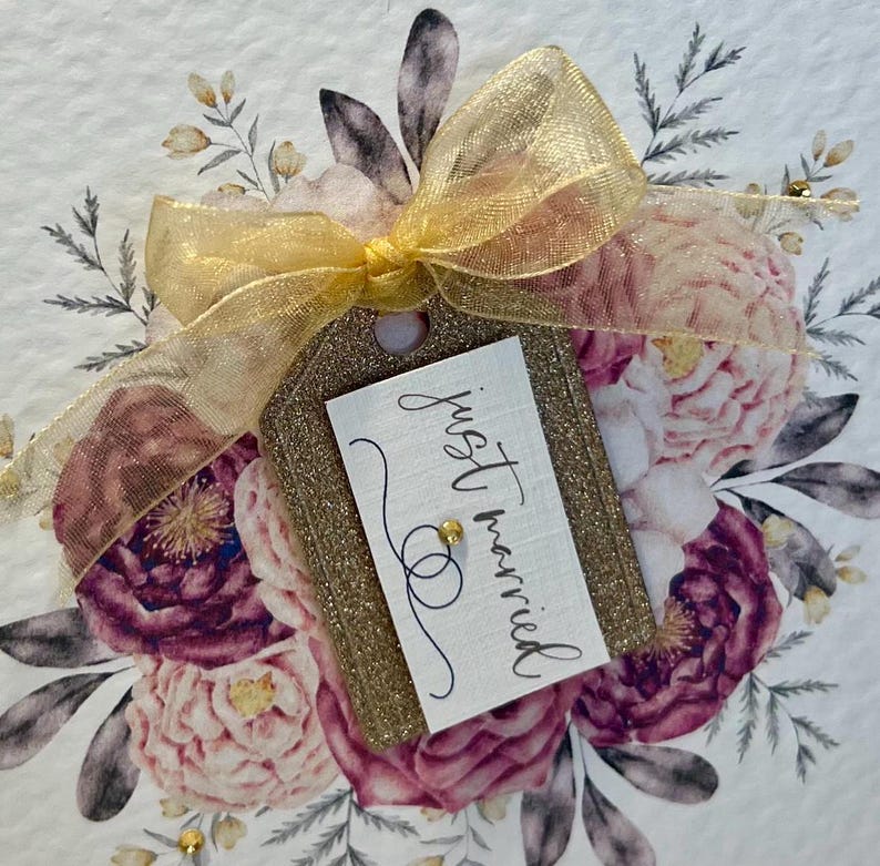 May include: A gold glitter tag with a white card that says "just married" in black calligraphy. The tag is tied with a gold ribbon and is surrounded by a floral arrangement of pink and purple flowers.